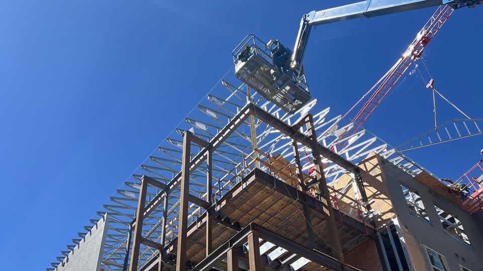 Sunlight over a mid rise building with its steel frame and roof trusses still exposed, a boom lift and tower crane working at the roof line